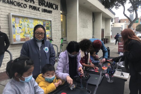 Kids and parents doing art and craft projects on a table in front of a library.