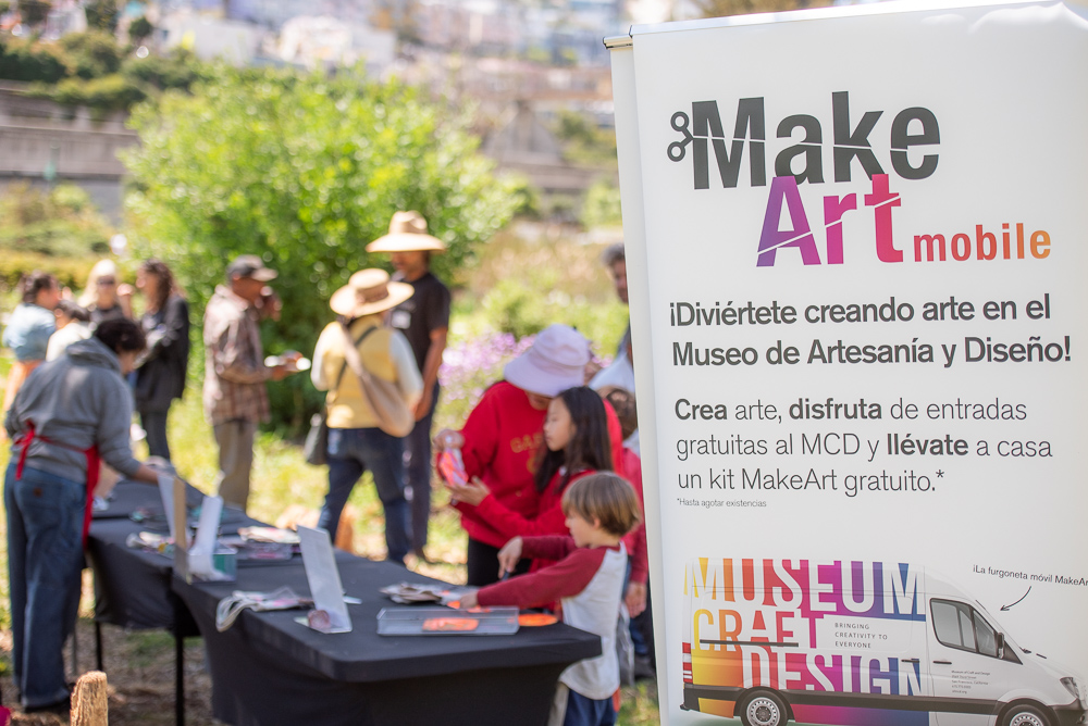 Photo of a MakeArt Mobile sign and people crafting at a table in the background.