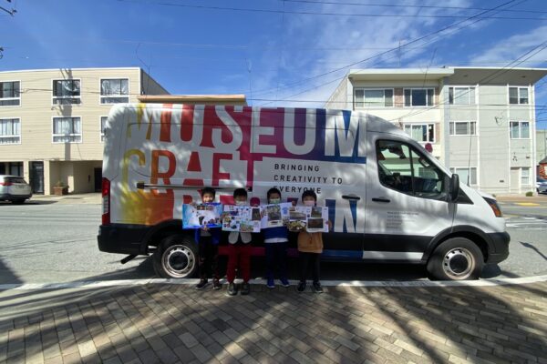 Four kids holding up their artwork standing in front of a van with the Museum of Craft and Design's logo on it.