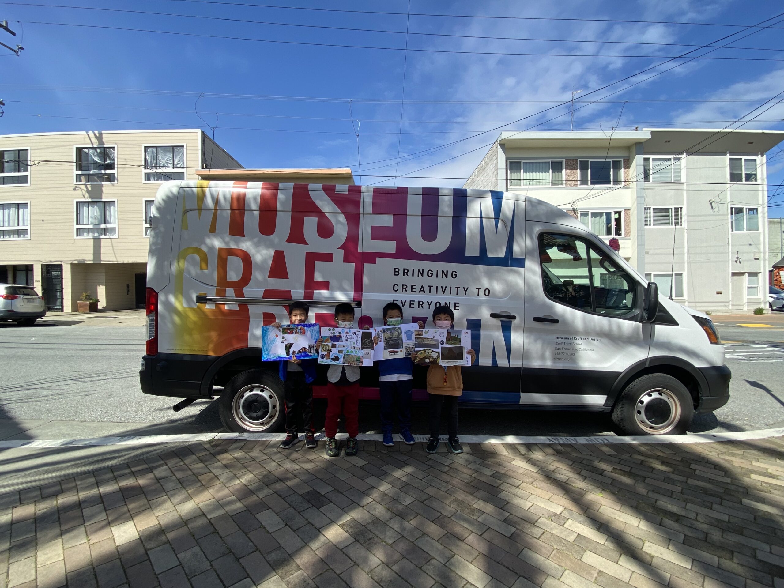 Four kids holding up their artwork standing in front of a van with the Museum of Craft and Design's logo on it.
