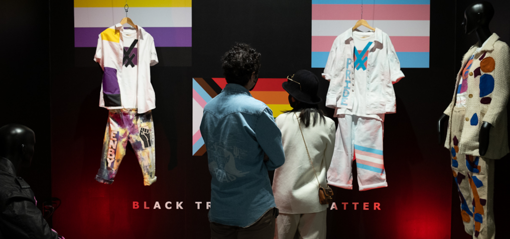 exhibition photo of outfits hanging in front of two people with their backs to the camera and multi-colored flags on the back wall