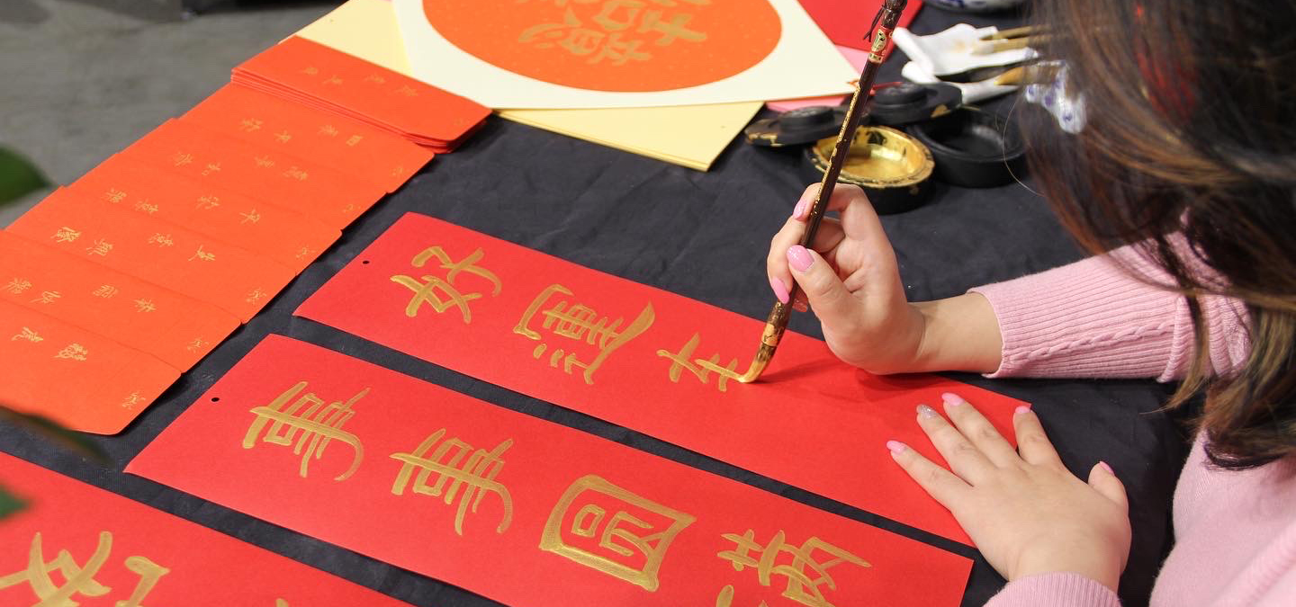 Person writing Chinese brush calligraphy in gold ink on a red paper