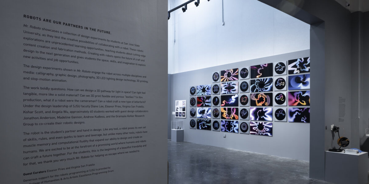 Photo of an art exhibition with text on the wall and two dimensional work on a wall in the background
