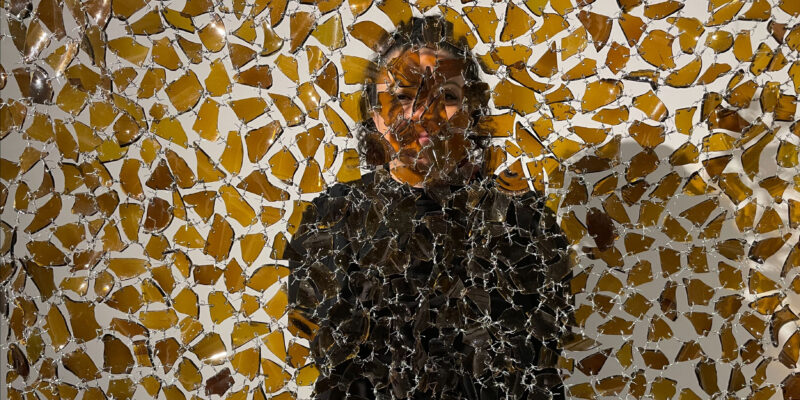 Artwork made up of hanging pieces of broken brown beer bottles with a person standing behind looking through.