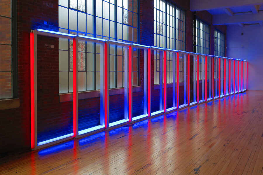 Photo of a neon artwork constructed out of rectangles that are red on the vertical and blue on the top, and standing on their own down a wooden floor.