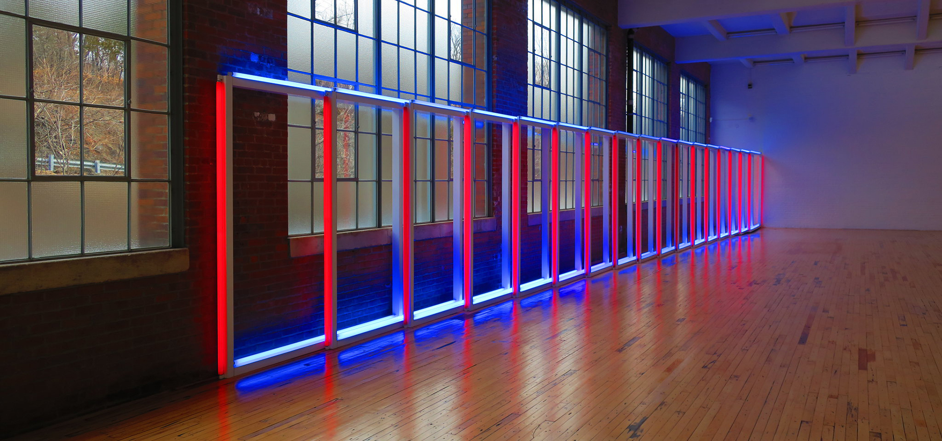 Photo of a neon artwork constructed out of rectangles that are red on the vertical and blue on the top, and standing on their own down a wooden floor.