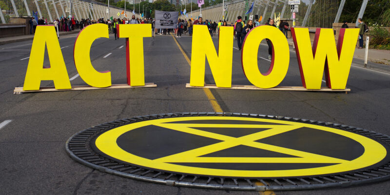 Photo of large yellow letters spelling out ACT NOW on a street with people in the distance behind it