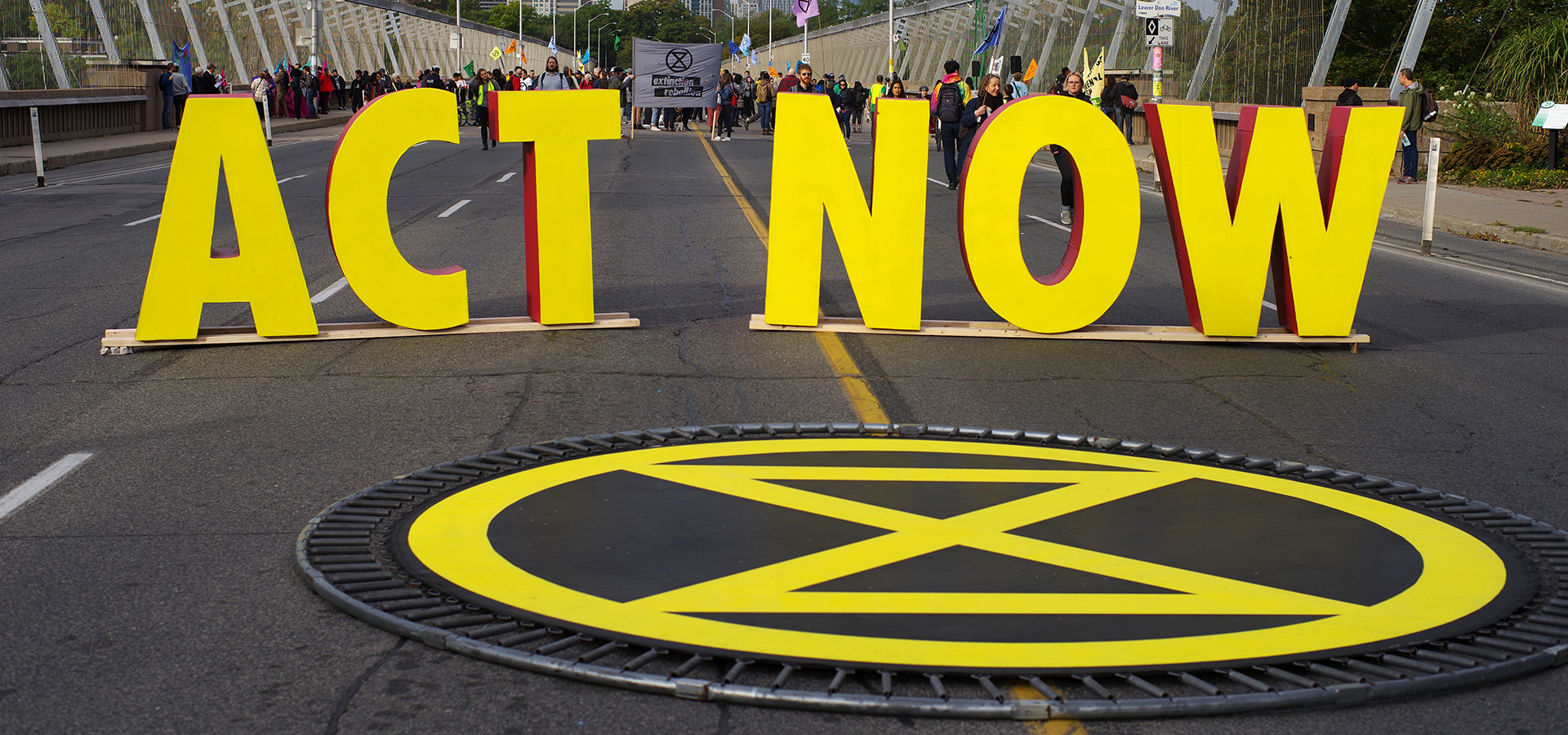 Photo of large yellow letters spelling out ACT NOW on a street with people in the distance behind it