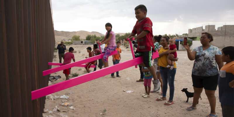 photo of kids sitting on one side of a pink teeter-totter that is connected a wall at the border. People are standing behind them