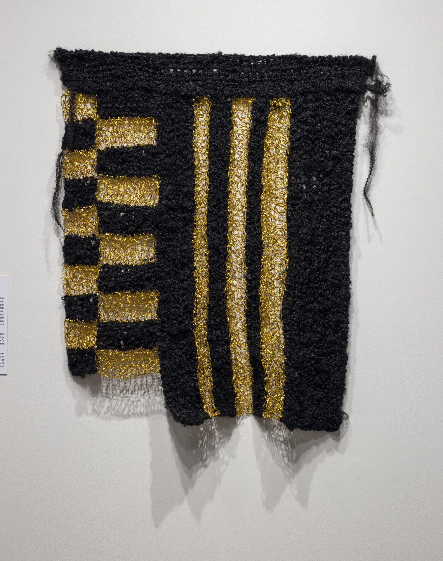 Artwork hanging on a wall. Black and gold textile to form vertical pattern.