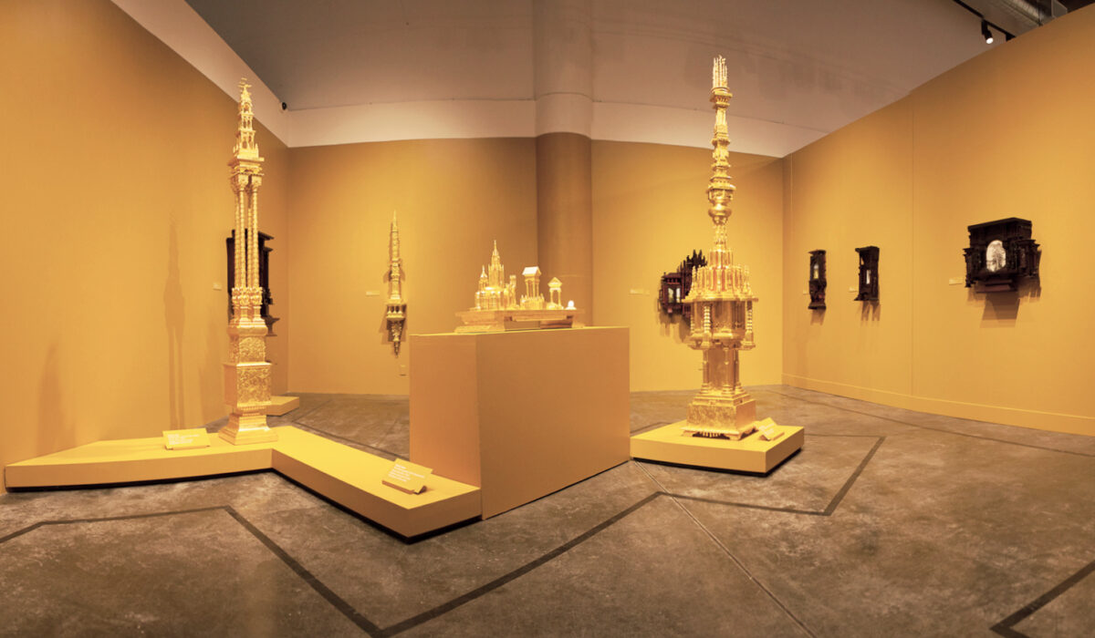 Panoramic photo of an exhibition. All walls and pedestals are yellowish gold with sculptures hanging on walls and placed on pedestals