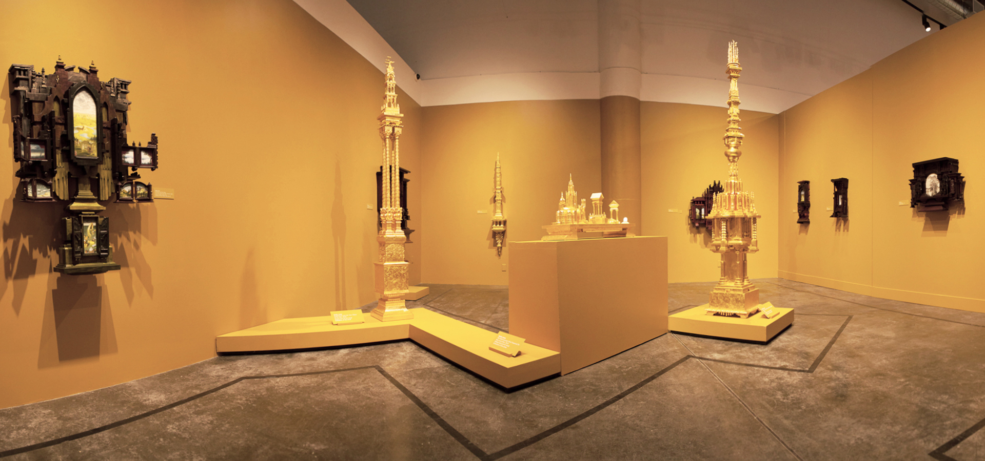 Panoramic photo of an exhibition. All walls and pedestals are yellowish gold with sculptures hanging on walls and placed on pedestals