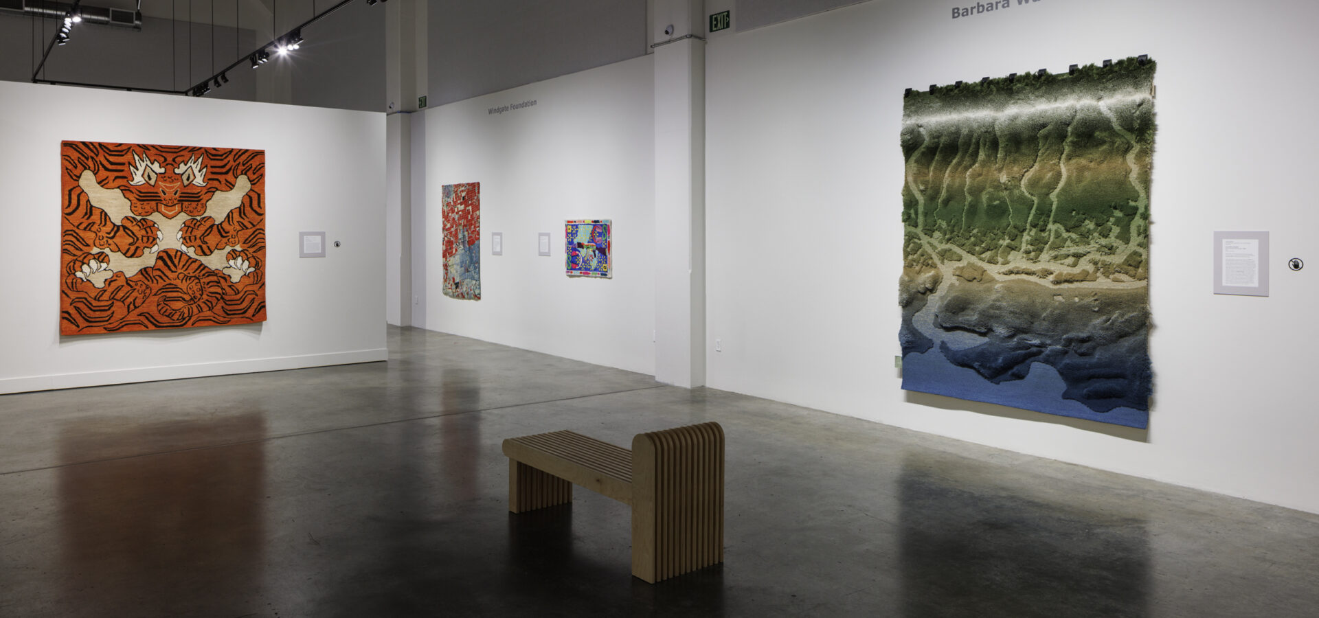 Photo inside a museum of an exhibition featuring rugs hanging on walls and on pedestals on the floor.