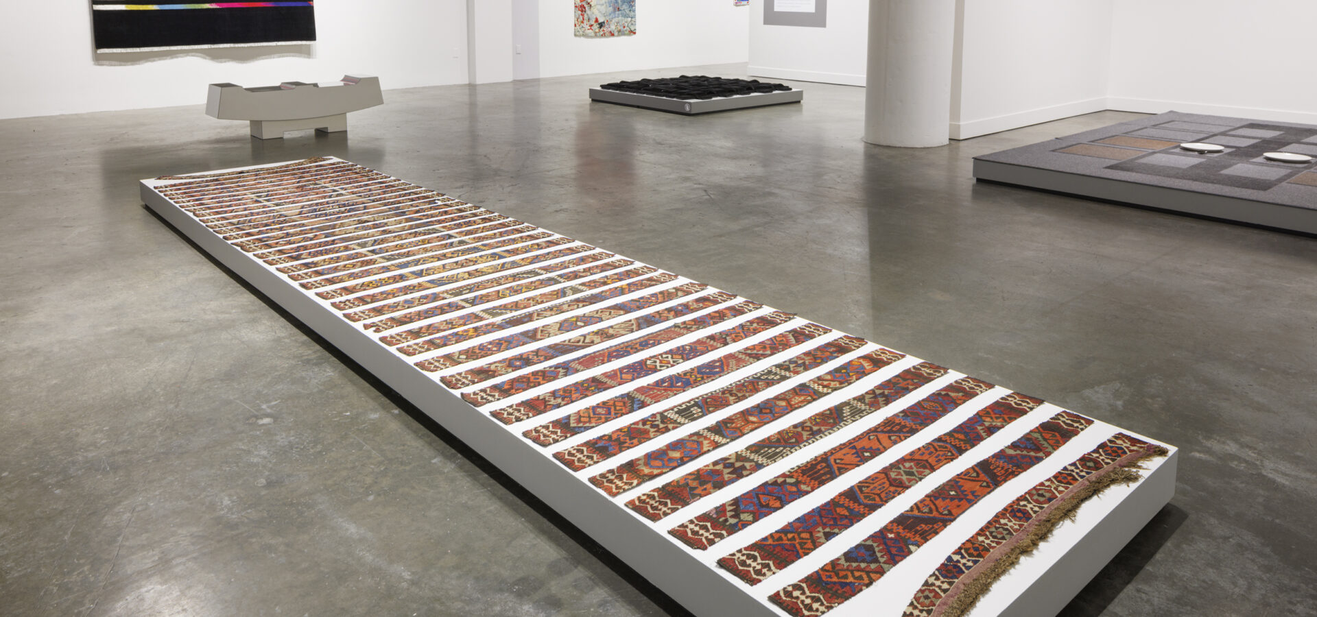 Photo inside a museum of an exhibition featuring rugs hanging on walls and on pedestals on the floor.