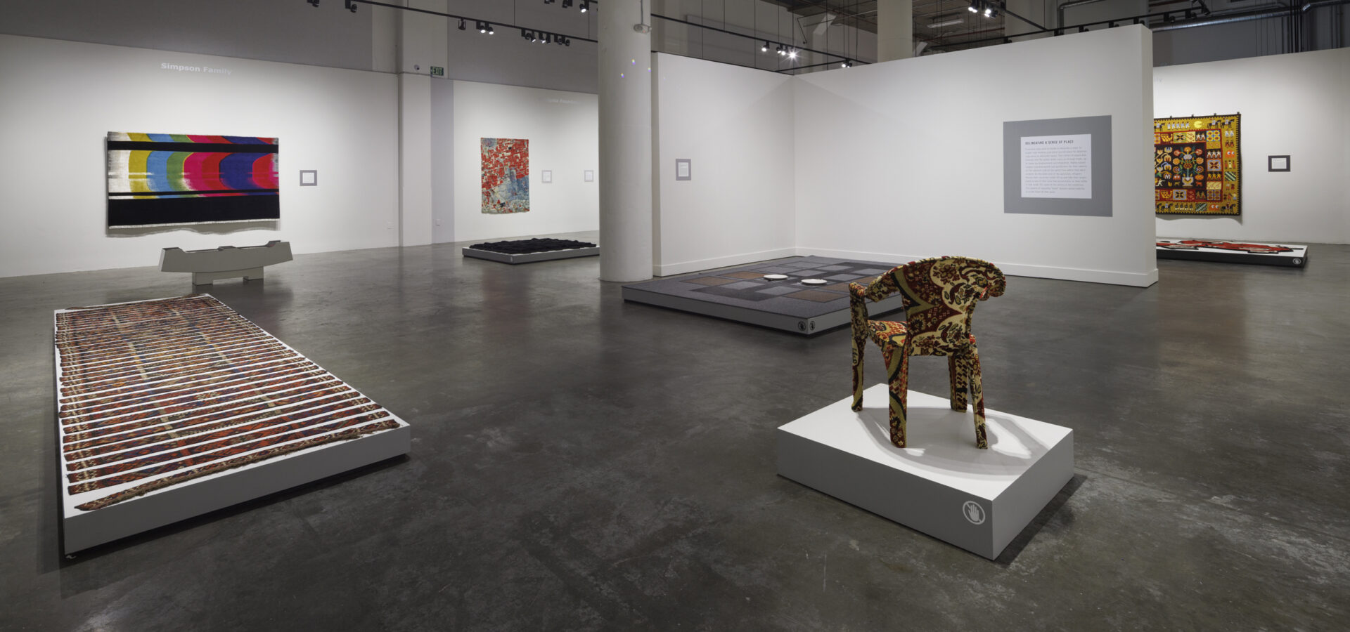 Photo inside a museum of an exhibition featuring rugs hanging on walls and on pedestals on the floor.