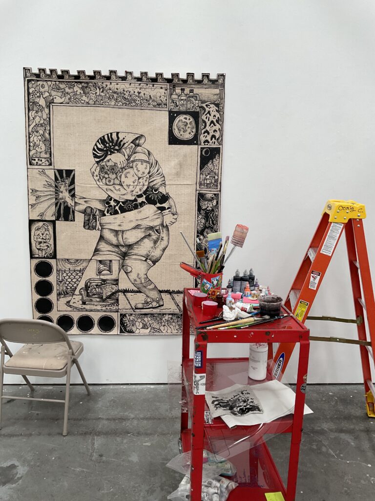 Photo of a monochromatic tapestry hanging. A metal folding chair sits and orange ladder in front of it with a red rolling cart/table filled with art supplies