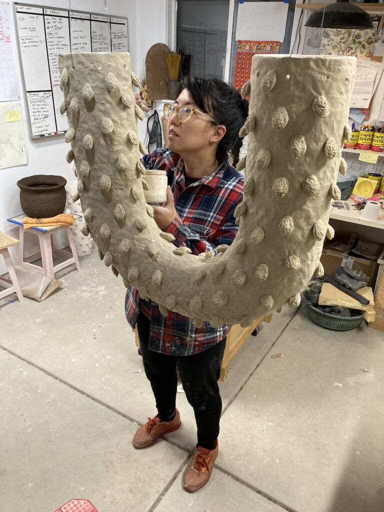 Photo of Cathy Lu (artist) in her studio with a hanging piece of ceramic art that is shaped in a U