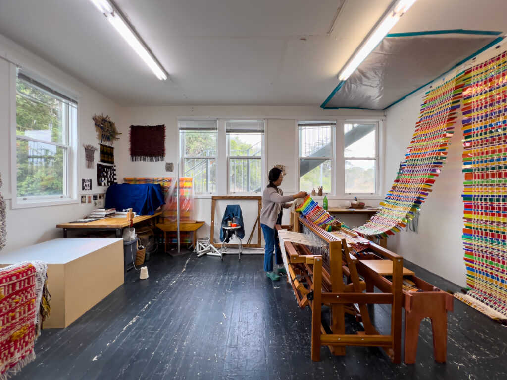 Photo of an artist holding a multi-color woven tapestry hanging on the wall behind a loom in her studio.