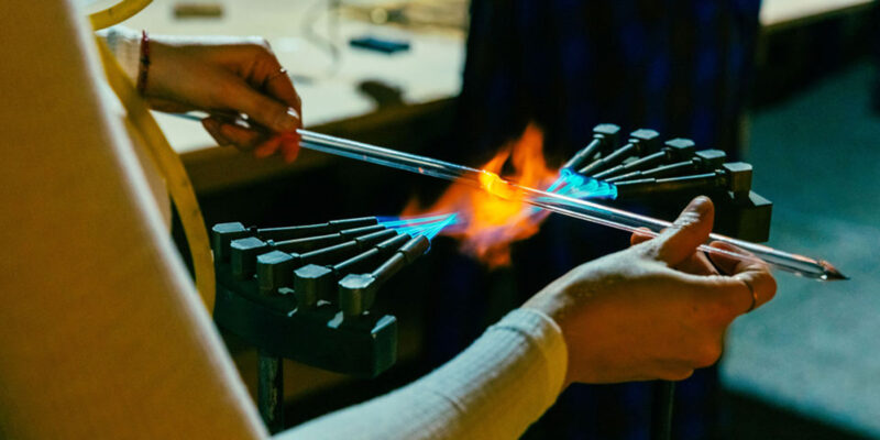 Close up photo of a person's hands holding a thin long piece of glass neon over fire to bend it.