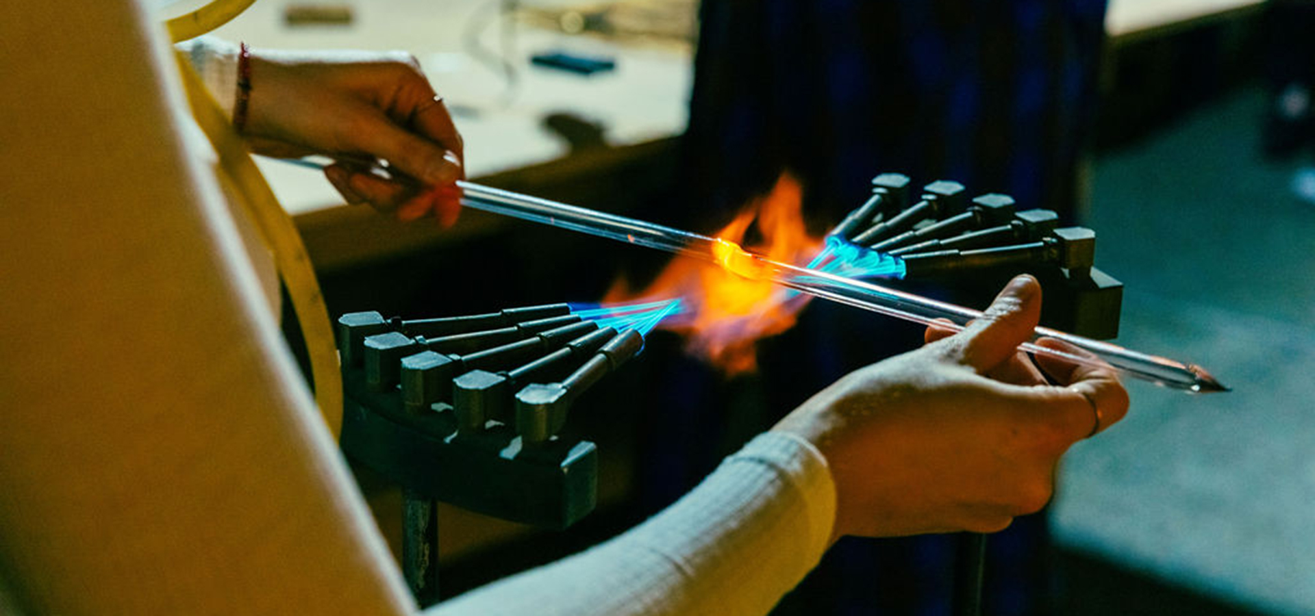 Close up photo of a person's hands holding a thin long piece of glass neon over fire to bend it.