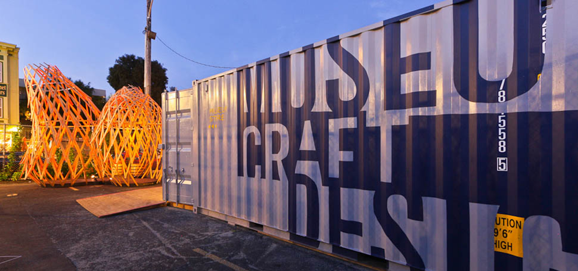 Three vertical wooden lattice structures with a shipping container in front of them with the Museum of Craft and Design's logo on it.