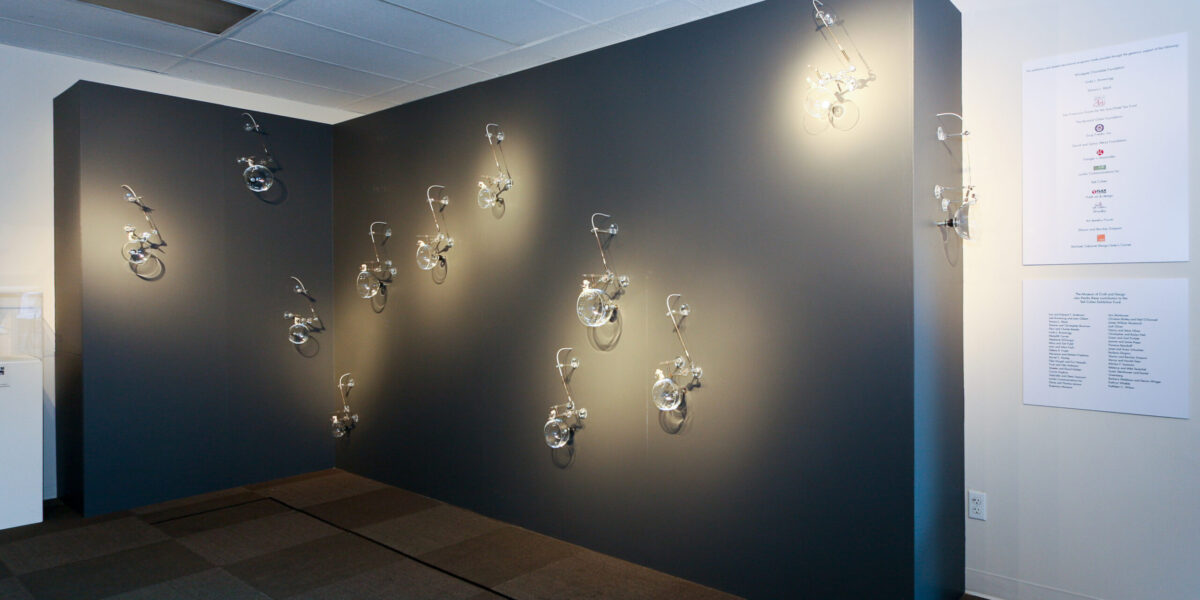 View of inside and exhibition with grey wall with hanging glass objects.
