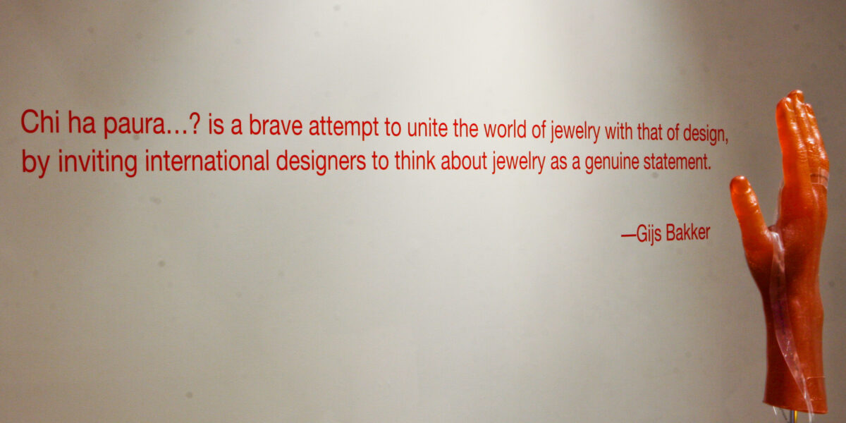 an exhibition photo of a gallery wall with a quote that reads, "Chi ha paura...? is a brave attempt to unite the world of jewelry with that of design, by inviting international designers to think about jewelry as a genuine statement." -Gis Bakker