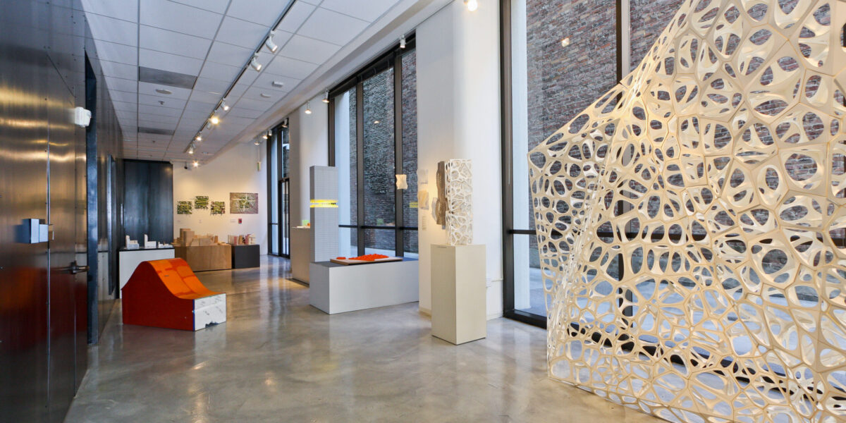 Photo of an exhibition with a large geometric white sculpture in front of tall windows.