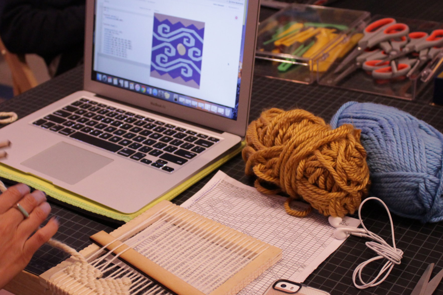 Photo of a pair of hands doing a weaving project in front of a laptop and weaving tools