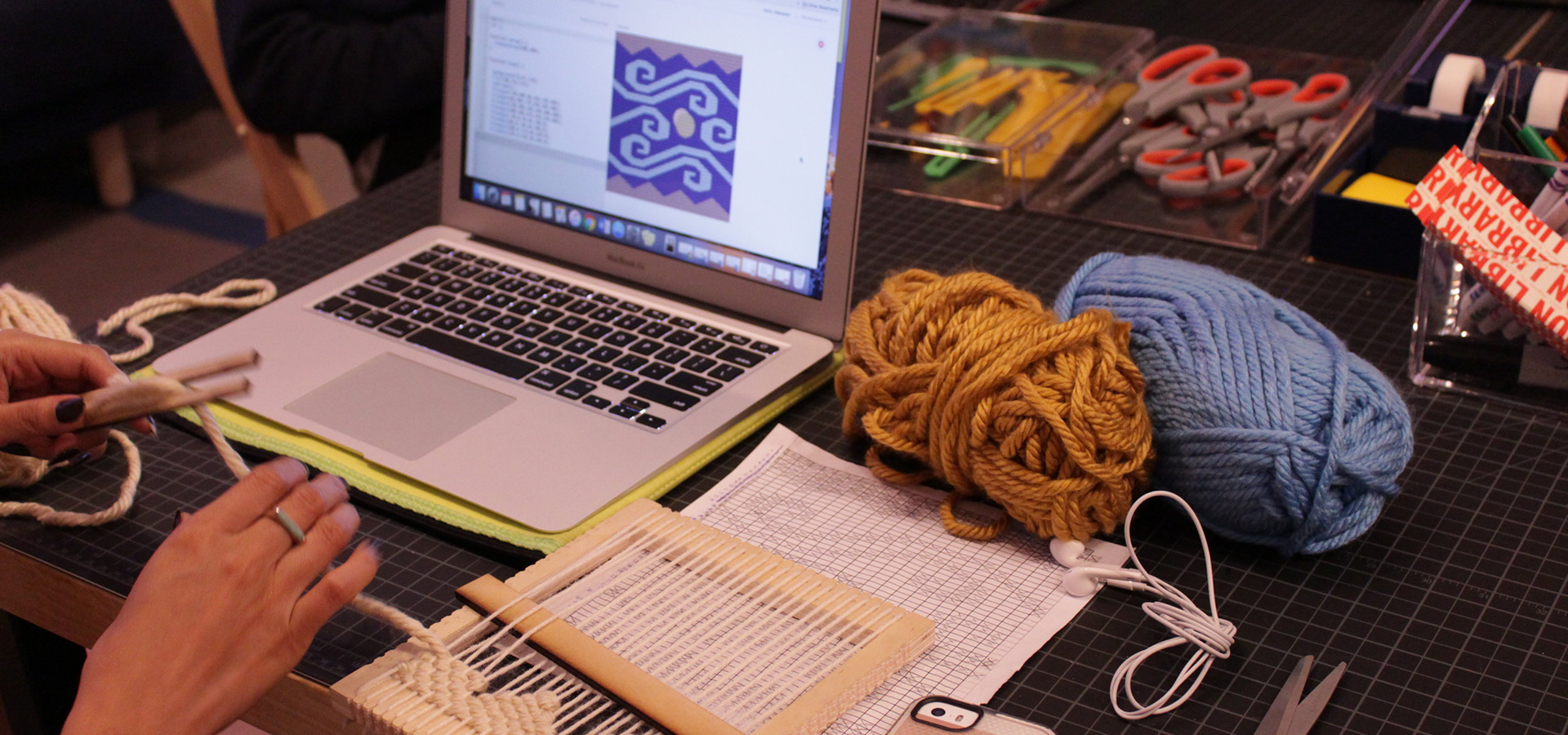 Photo of a pair of hands doing a weaving project in front of a laptop and weaving tools