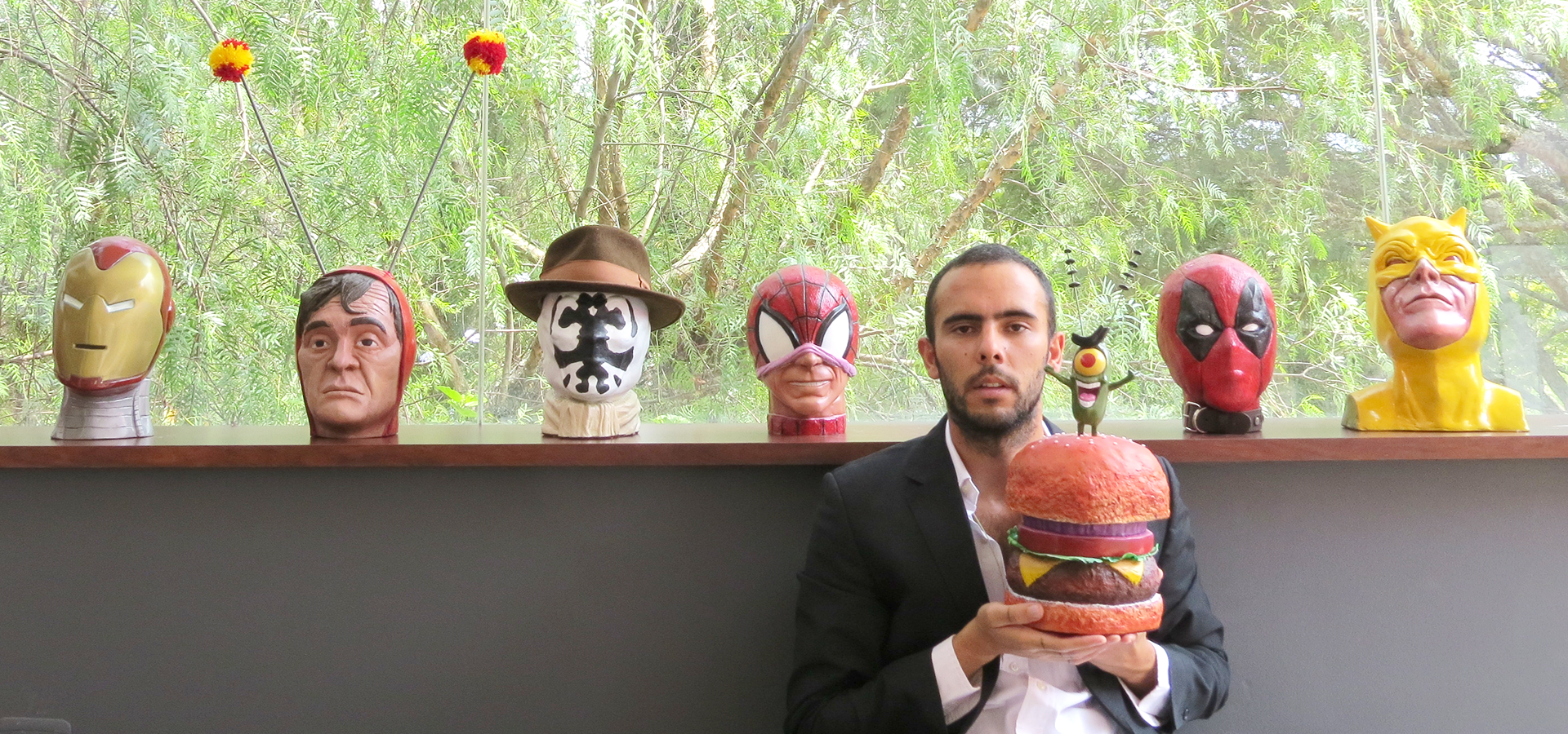 Headshot of Daniel Iturralde helding a plankton and kraby patty sculpture with his papier-mâché head sculptures of comic and cartoon characters, including iron man, spiderman, and others.