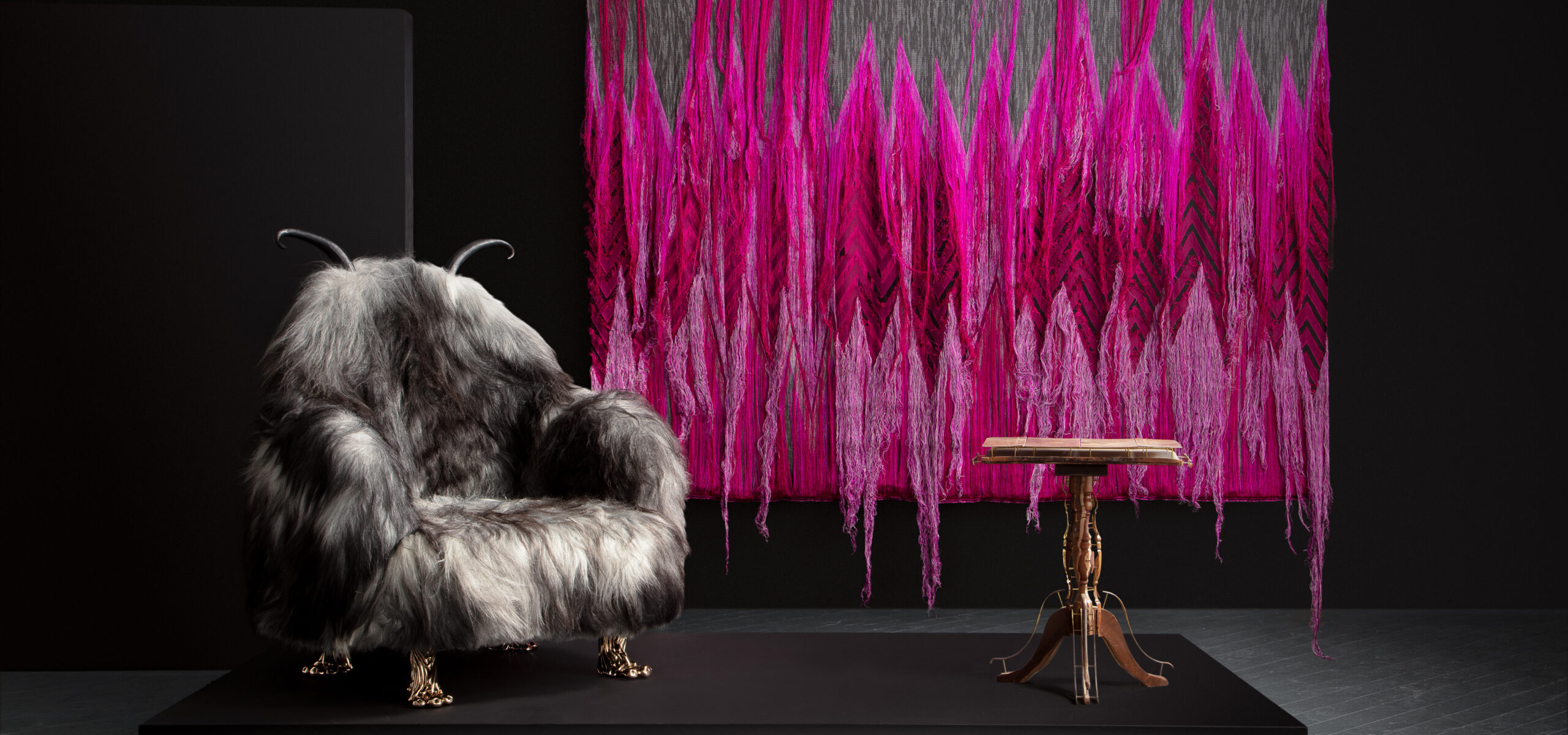 Photo of an exhibition with a hanging dark pink fiber object, fur like covered chair and wooden table