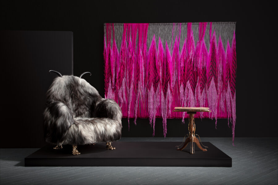 Photo of an exhibition with a hanging dark pink fiber object, fur like covered chair and wooden table