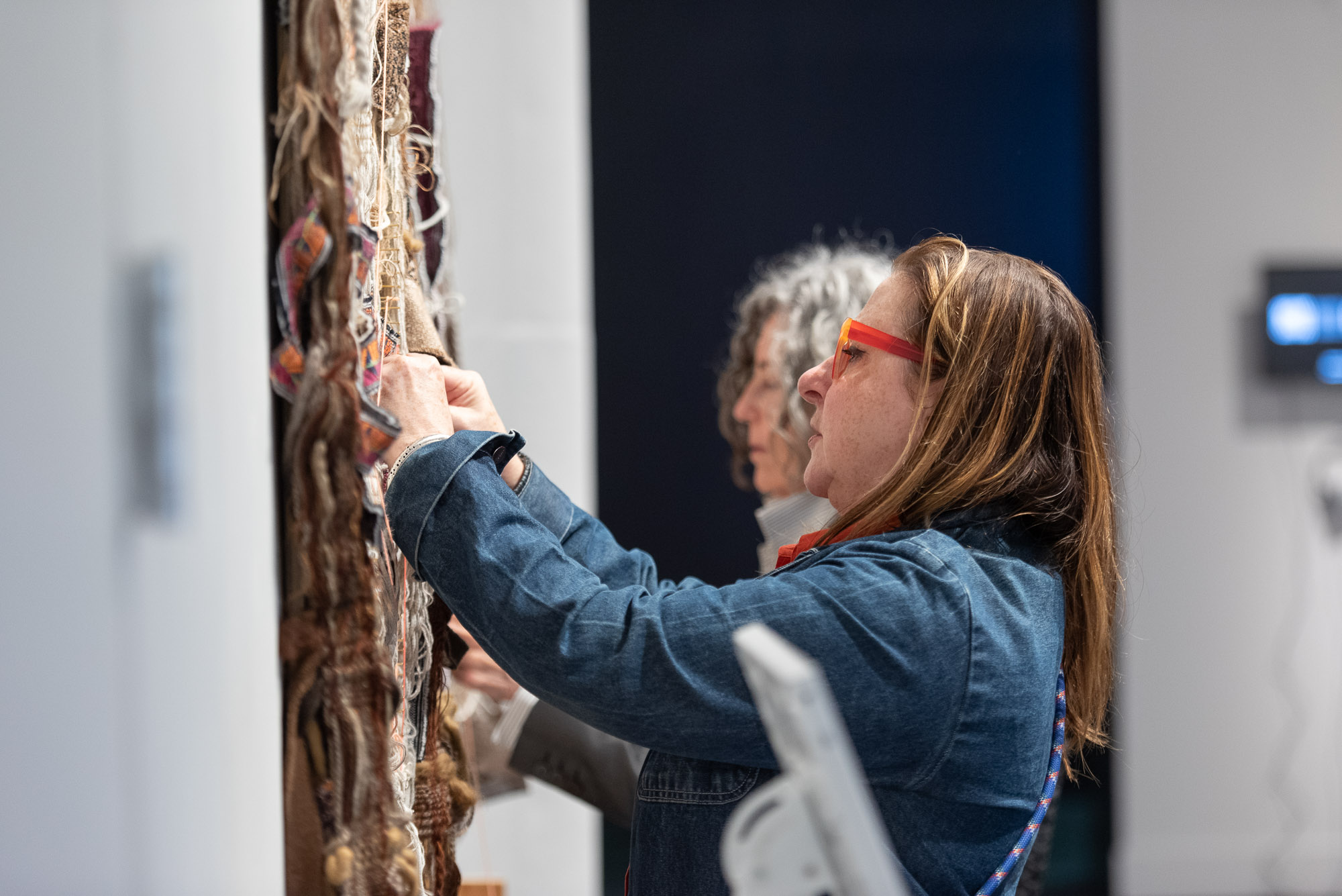 Two people threading yarn on artwork hanging from the wall.
